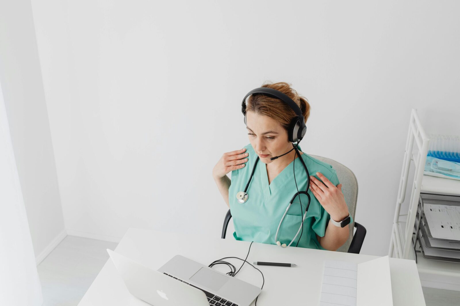 Female doctor in scrub suit conducting an online consultation using a laptop and headset in a bright office space.